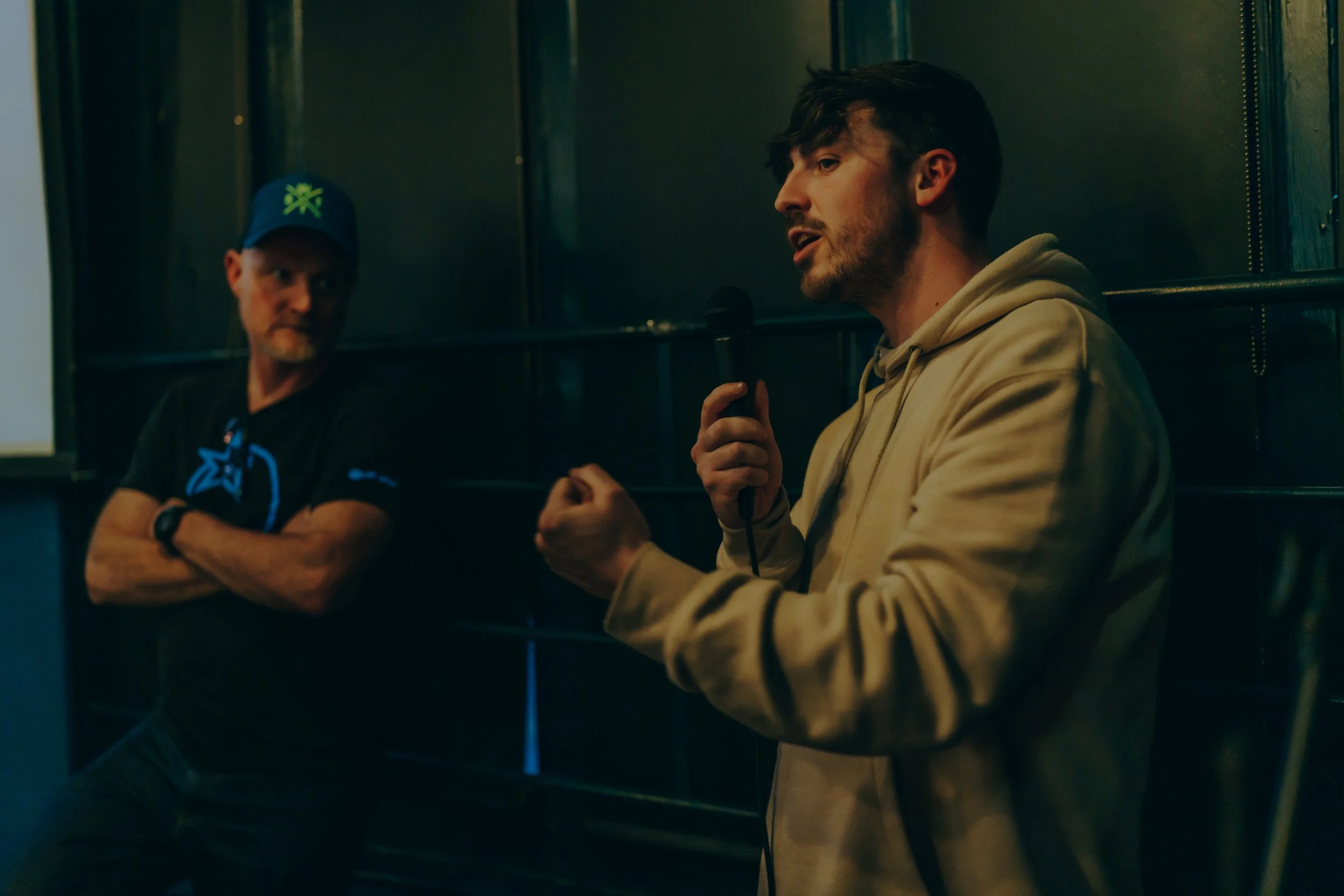 Man in a beige hoodie holding a microphone and speaking while another man in a black t-shirt and blue cap watches attentively.