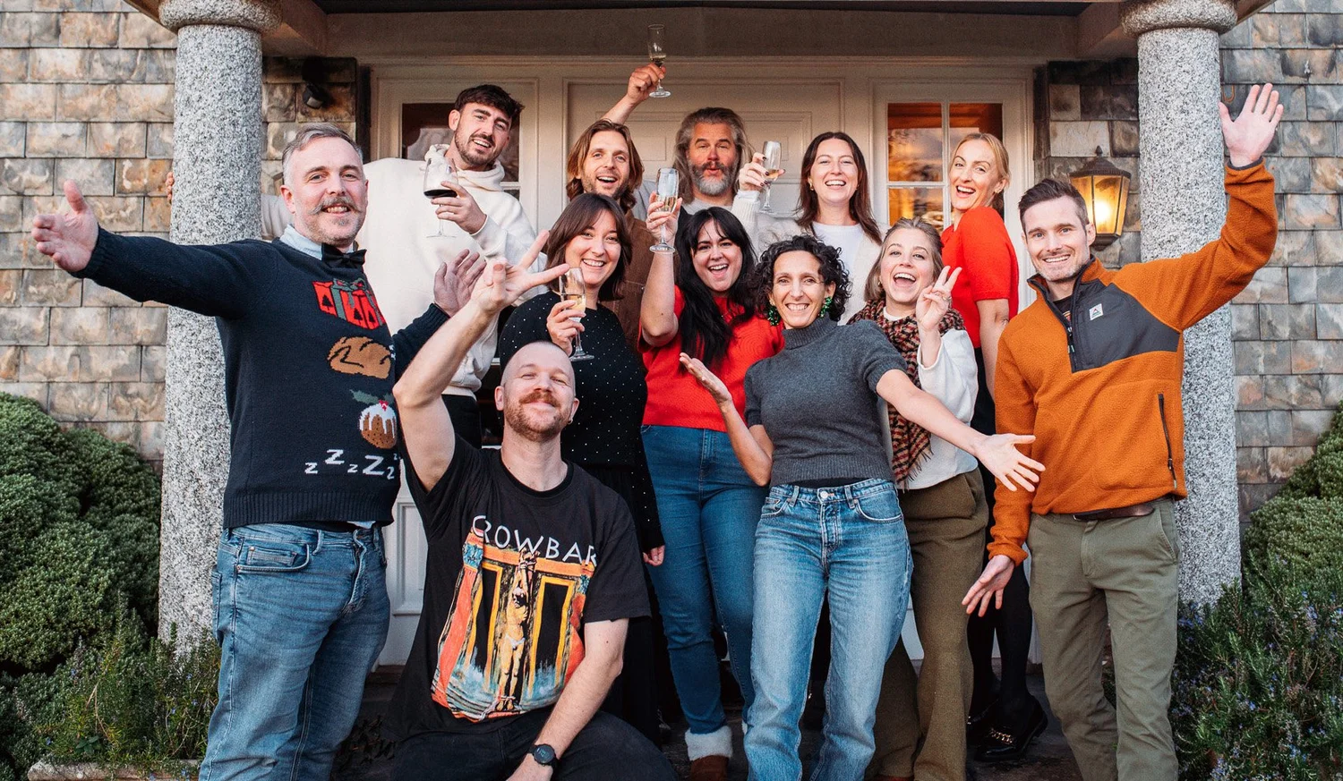 Group of twelve adults smiling and raising glasses, posing happily outside a stone building entrance.