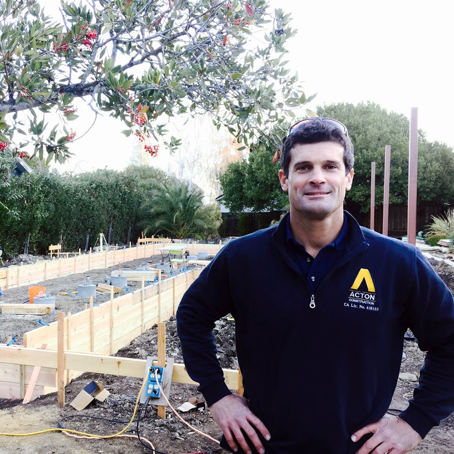 Man in a navy blue jacket with Acton Construction logo standing with hands on hips at a construction site with wooden framing and greenery in the background.