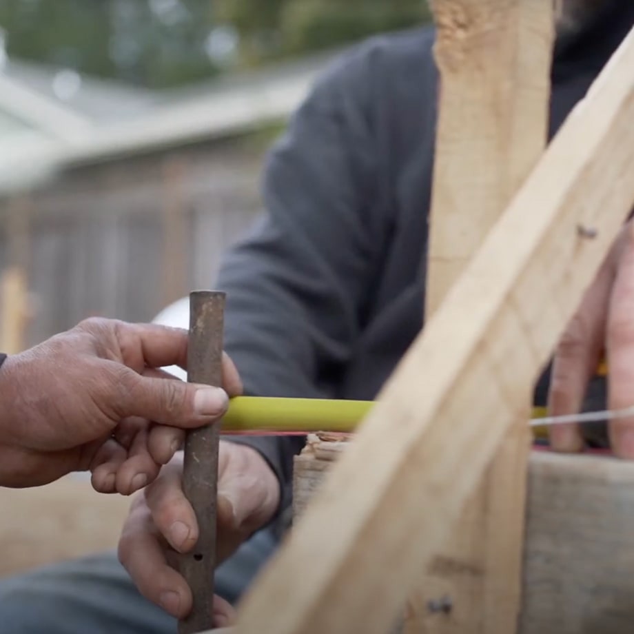 Two hands holding a rusted metal rod and a yellow measuring tape against wooden construction materials.
