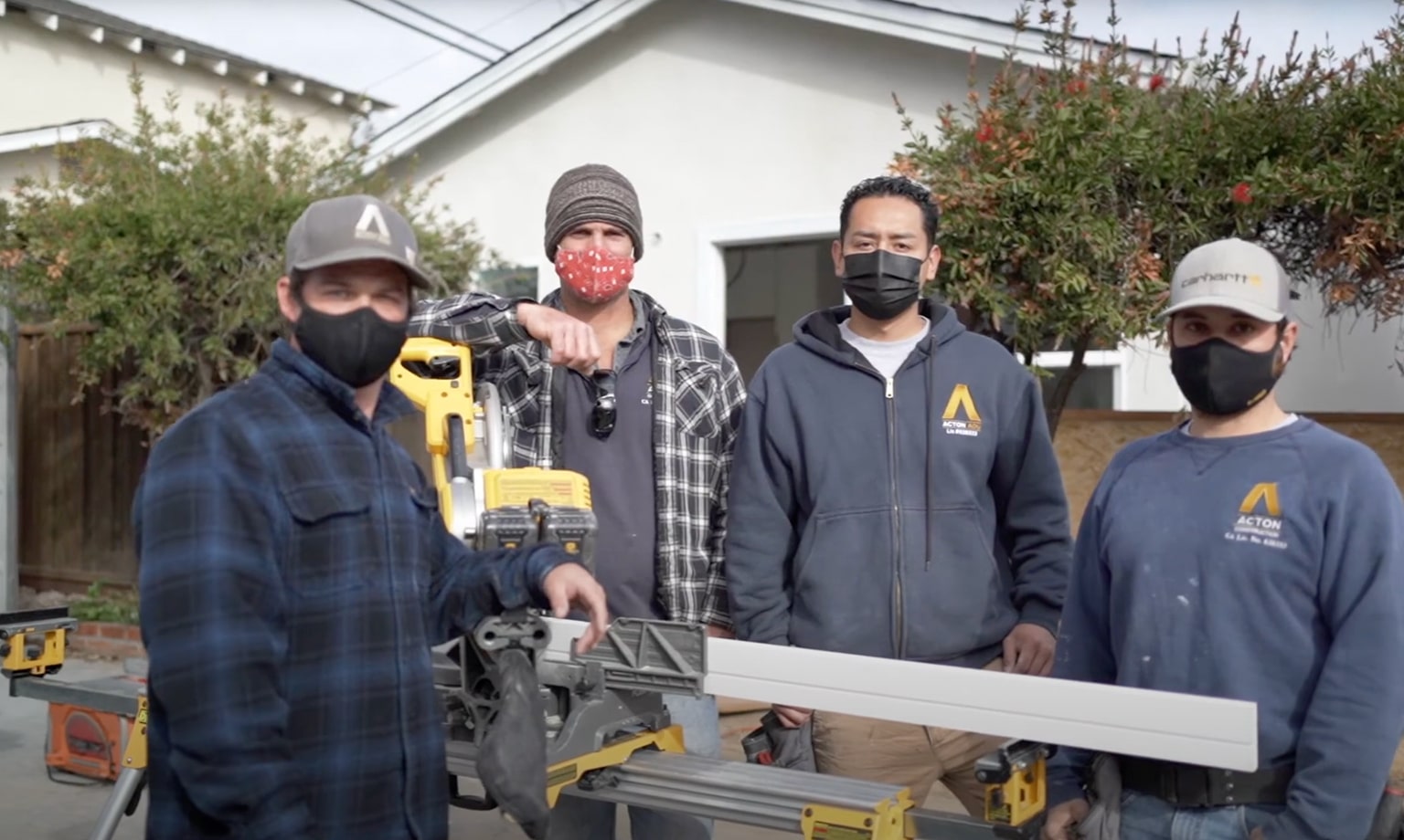 Four construction workers wearing masks standing around a yellow miter saw outdoors in front of a house.