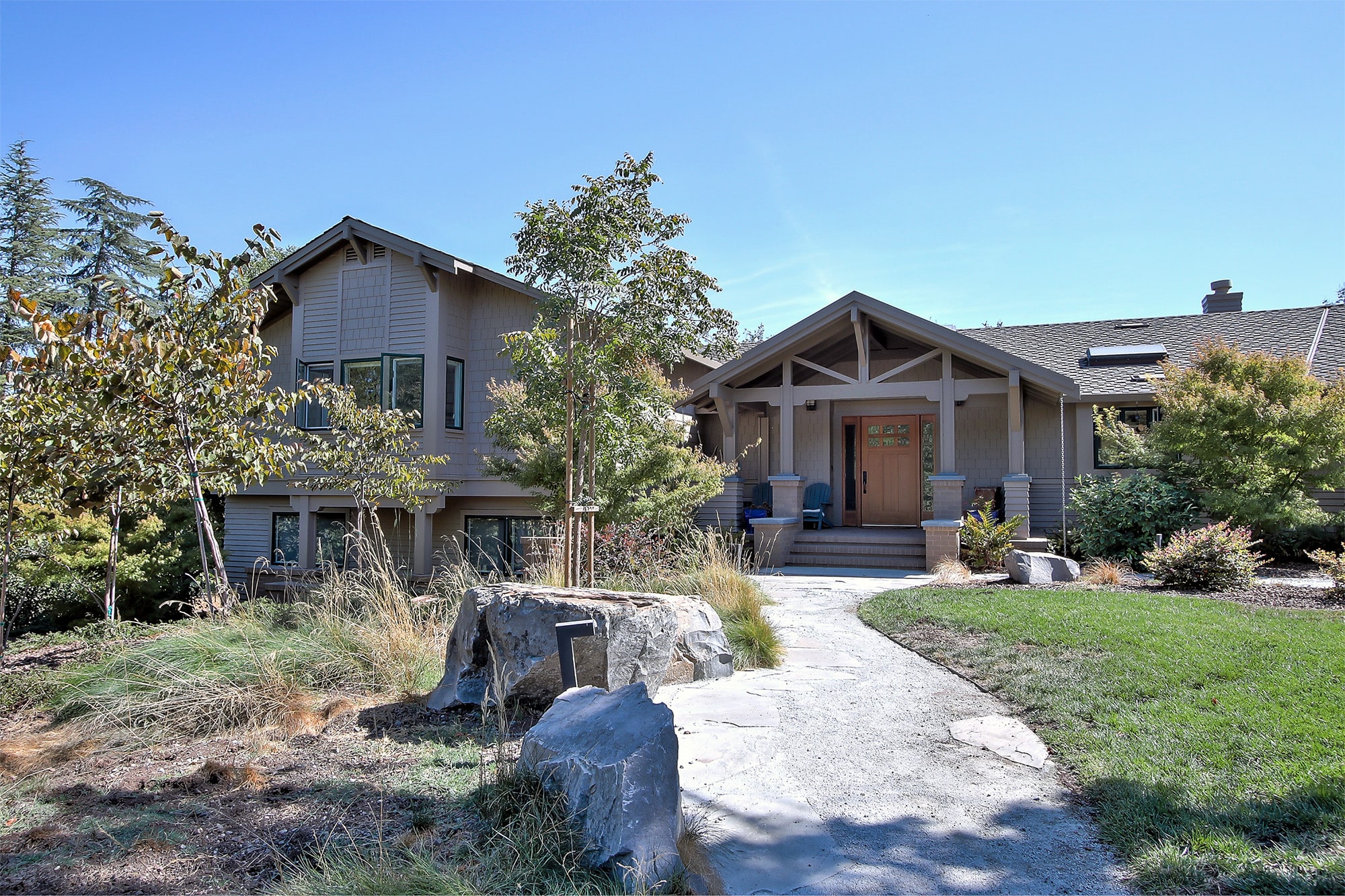 Front view of a modern single-story house with beige siding, wooden door, and landscaped yard with trees and rocks.