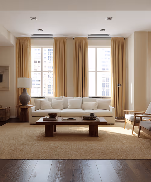A modern living room featuring two large windows, minimalist design, and warm wooden floors and furniture, highlighting a seamless blend of comfort and architectural elegance.