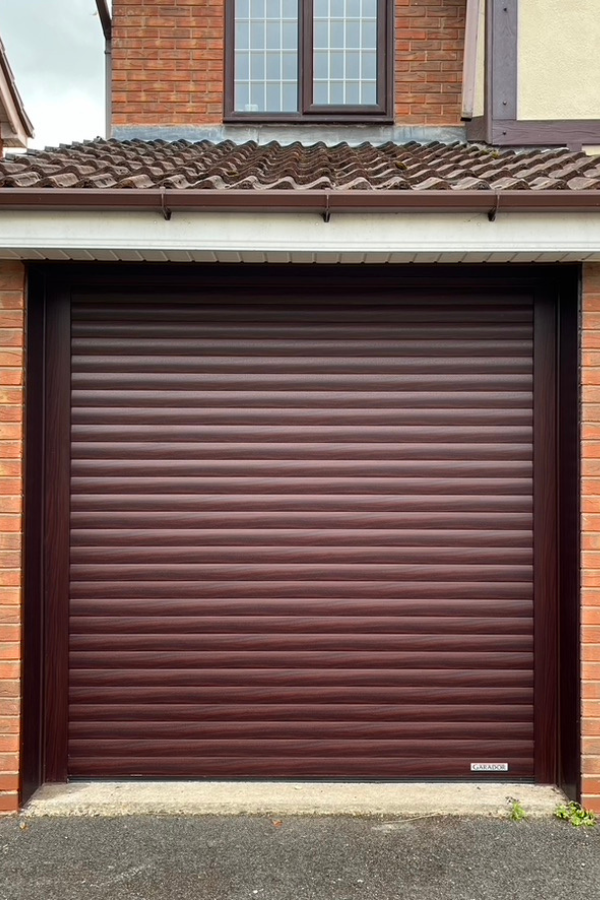 Closed brown horizontal slatted garage door set in a brick house facade with a tiled roof and a window above.