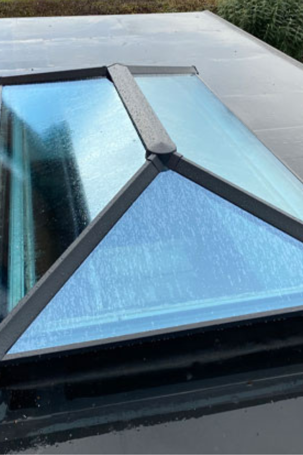 Triangular glass skylight with black metal frame on a flat roof, with water droplets on the glass.