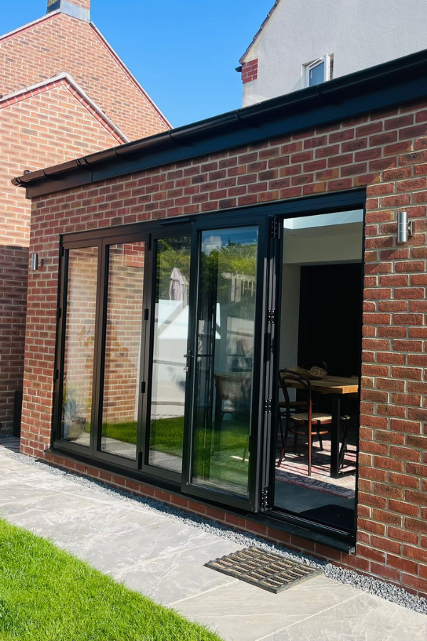Modern black-framed bifold glass doors open to a dining area in a brick house with a stone patio and green lawn outside.