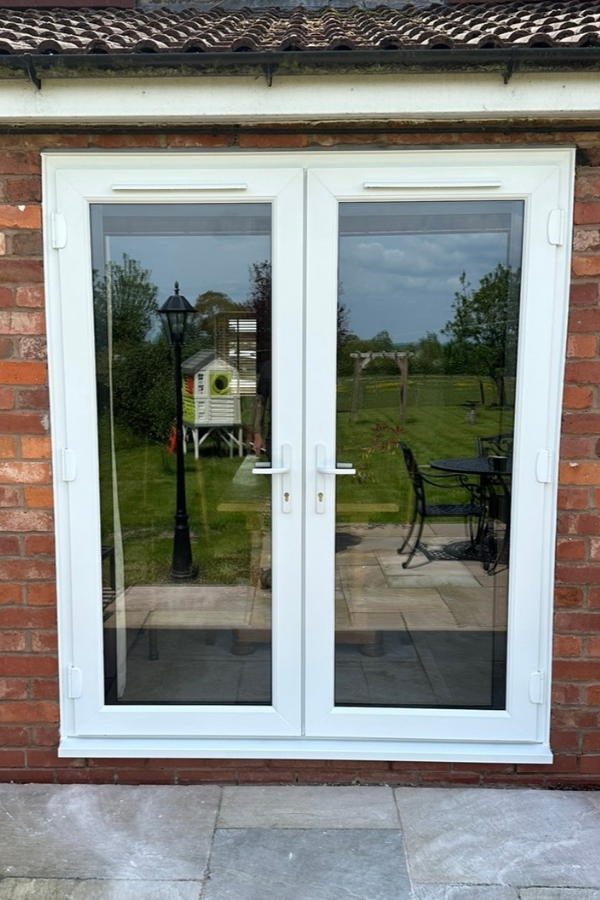 White framed double glass French doors opening to a garden with a lawn, birdhouse, black lamp post, and patio furniture.