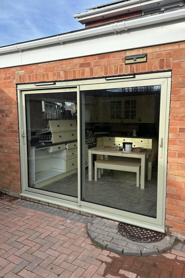 Large beige sliding patio doors on a brick house, revealing a kitchen with yellow cabinets and a wooden table with a bench inside.