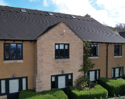 Two-story beige residential building with multiple black-framed windows, green bushes, and a sloped roof with skylights.