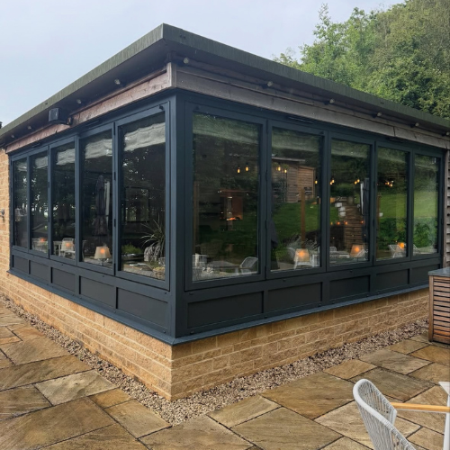 Corner view of a modern glass-enclosed sunroom with black frames on a stone patio beside a grassy area.