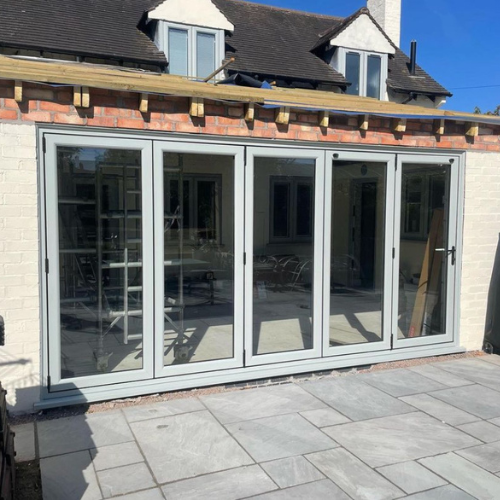 Modern green four-panel bifold glass doors installed on a white brick house with a stone patio in front.