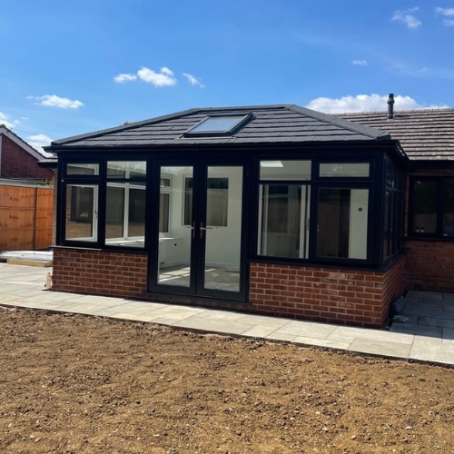 New brown Edwardian-style conservatory with black framed windows and double doors on a brick base, attached to a house under a blue sky.