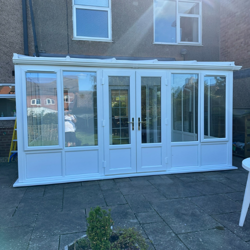 White lean-to conservatory attached to a brick house with glass double doors and multiple windows.