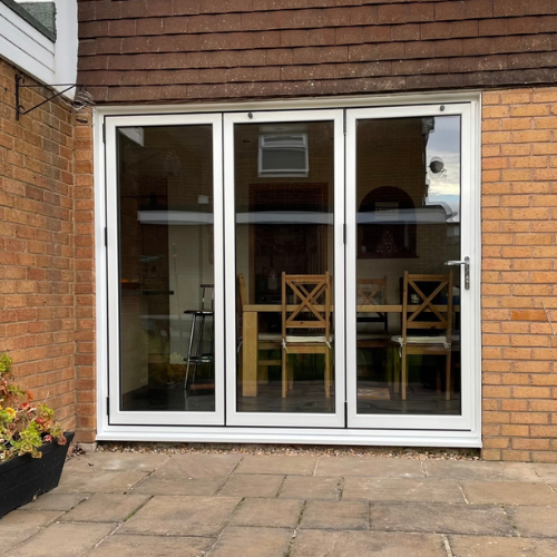 White-framed glass bifold doors on a brick house with a view of wooden chairs and a table inside.