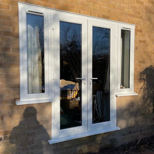 White-framed French doors with glass panels installed in a brick wall, showing reflections of trees and a shadow of a person in front.