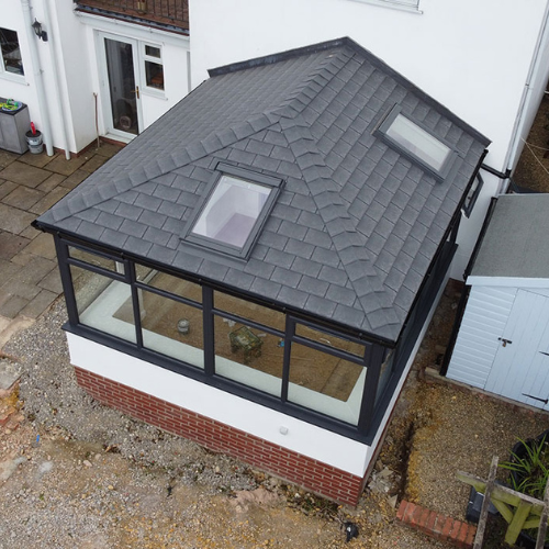 Aerial view of a modern conservatory with a grey tiled pitched roof with two skylights, large black-framed windows, and red brick base attached to a white house.