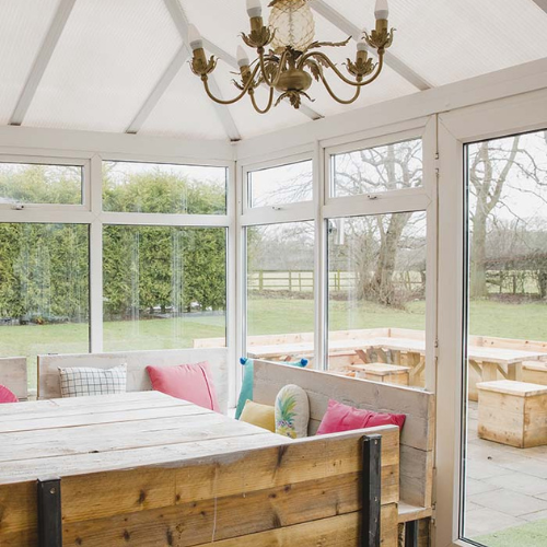 Interior of a bright Victorian conservatory with wooden seating, colourful cushions, a wooden table, and a brass chandelier hanging from a glass roof.