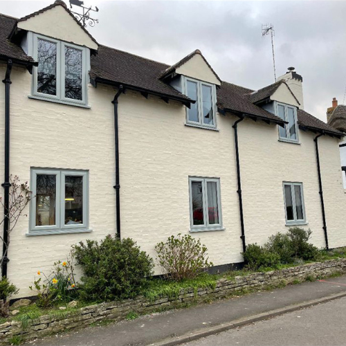 White brick house with brown tiled roof, six uPVC windows, and garden shrubs alongside a stone border.