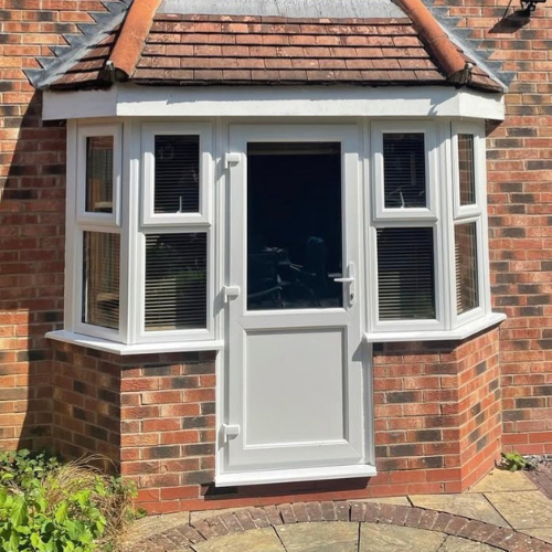 White UPVC door with glass panel surrounded by white framed bay windows on a red brick house.