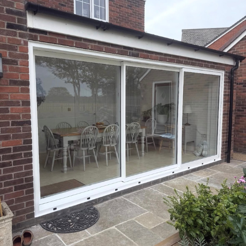 White-framed aluminium sliding patio door on a brick house showing a dining table with eight chairs inside.
