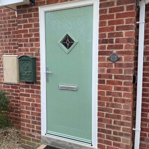 Pale green composite front door with a diamond-shaped window and silver handle, set in a red brick wall with a green mailbox and a beige utility box to the left.