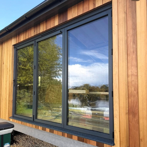 Modern black-framed glass bifold door on a wooden exterior wall reflecting trees and sky.