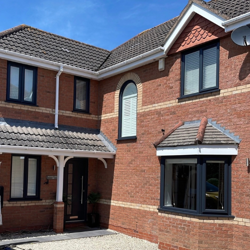 Two-story brick house with tiled roof, black-framed aluminium windows, and a covered entrance under a blue sky.
