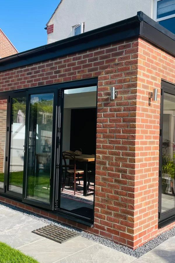 Modern orangery extension with red brick walls, large black-framed glass doors, and a view of a wooden dining table with chairs inside.