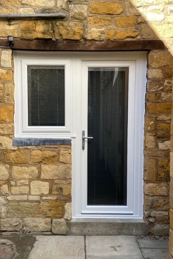 White uPVC door with vertical blinds next to a matching window with horizontal blinds, set in a stone wall.