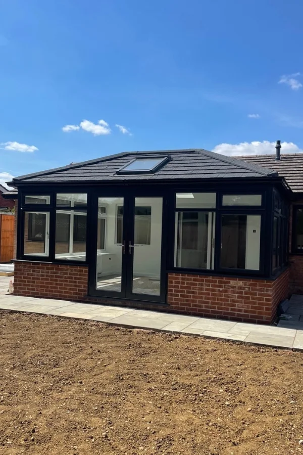 Modern conservatory with black frame, pitched roof, skylight, and brick base attached to a house under a clear blue sky.