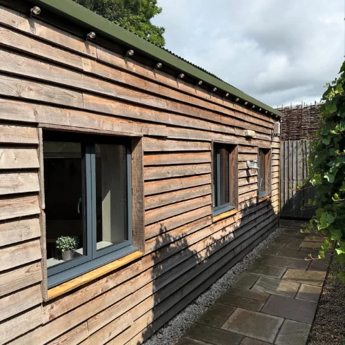 Exterior of a wooden building with three aluminum casement windows and a paved walkway beside it.