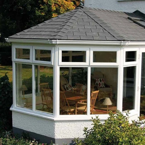 White uPVC-framed conservatory with glass walls and a grey tiled pitched roof, furnished with wooden chairs and a table inside.