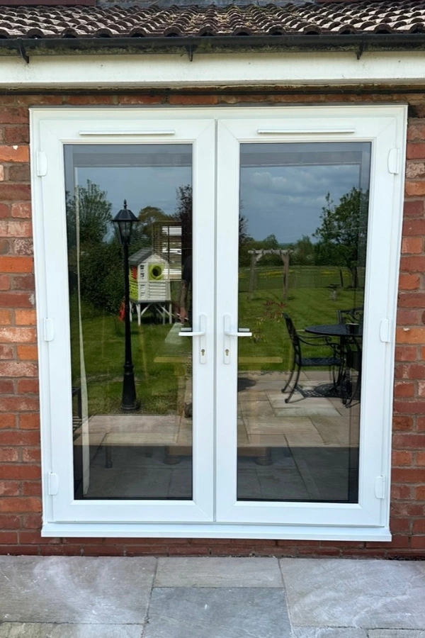 White uPVC double French doors with glass panes set in a brick wall, reflecting a garden with a lamp post, birdhouse, and outdoor furniture.