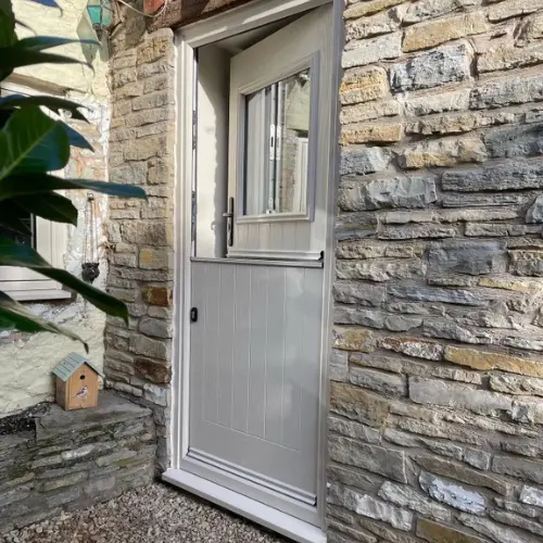 White composite stable door with a glass window in a rustic stone wall exterior with a small wooden birdhouse on a stone ledge nearby.