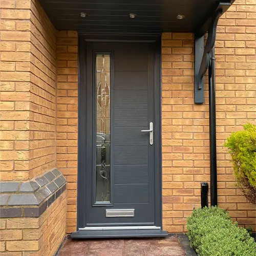 Dark grey composite front door with vertical glass panel and silver handle, set in a brick wall with plants nearby.