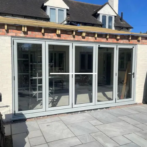 Light green uPVC bifold doors installed on a brick wall opening, leading to a patio with stone tiles under clear blue sky.
