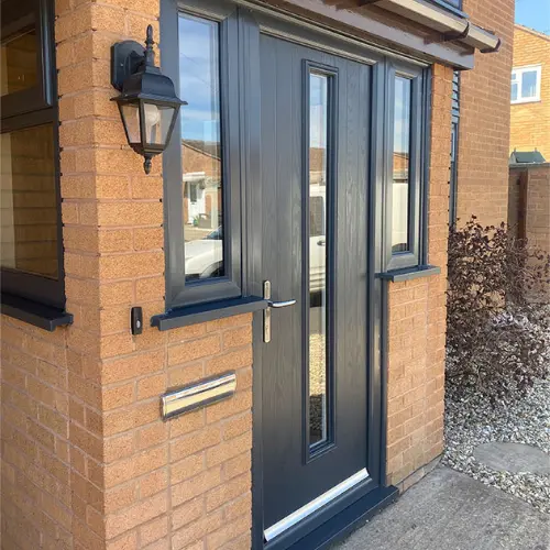 Modern dark gray composite front door with glass panels, mounted on a brick porch wall with a mail slot and a black lantern light.