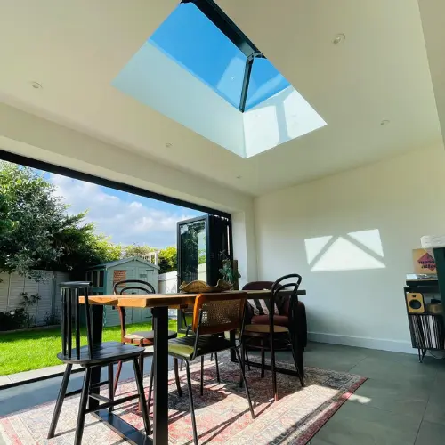 Modern orangery dining area with mixed chairs around a wooden table, featuring an open wall to a garden and a large skypod overhead.