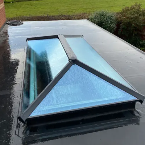 Rectangular glass skypod with a peaked frame installed on a flat black roof with grass and bushes in the background.
