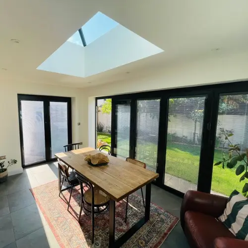 Modern orangery dining area with a wooden table and chairs under a large skylight, surrounded by black-framed glass doors opening to a sunny garden.