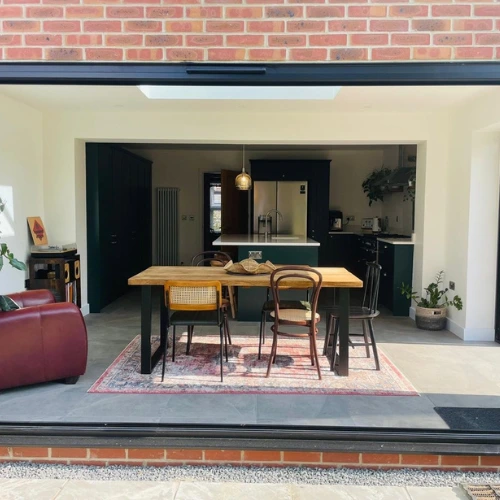 Modern orangery with an open kitchen with dark cabinets, a wooden dining table with mixed chairs on a rug, and a red armchair to the left.