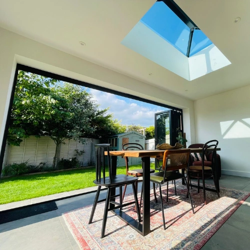 Modern orangery dining area with wooden table, mixed chairs on a rug, large bi-fold glass doors open to a sunny garden and a skylight above.