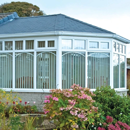 White Victorian conservatory extension with a tiled warm roof and vertical blinds inside, surrounded by a garden with flowering shrubs.