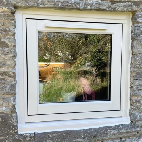 White framed square flush uPVC window set in a stone wall with reflections of trees and a pink flamingo garden ornament visible.