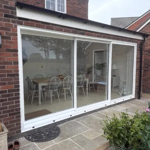 White aluminium sliding patio door opening to a dining area with a wooden table and eight chairs in a brick house.