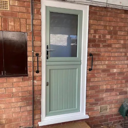 Green stable door with a frosted glass upper panel and vertical slats on the lower panel set in a white frame on a brick wall.