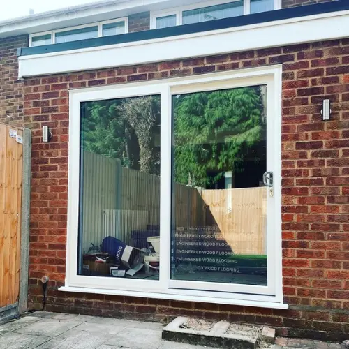 White uPVC sliding patio door installed on a brick exterior wall of a house with a wooden fence on the left and garden reflection on the glass.