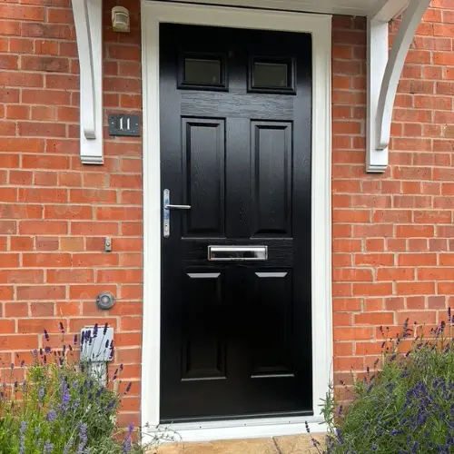 Black residential composite front door with silver handle and mail slot, set in a red brick wall with plants in front.
