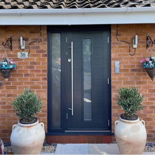 Modern dark grey front door with vertical glass panel, flanked by two large cream pots with green shrubs, on a brick house wall with hanging flower baskets and house number 36.