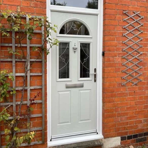 White front door with decorative glass panels set in a red brick wall, flanked by wooden trellises with climbing plants.
